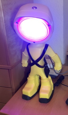 Yellow super-hero figurine in A-pose with tractor lamp head as desk lamp, shining red/blue RGB light.